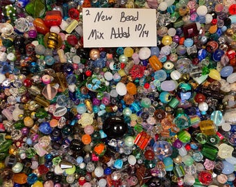 Puede incluir: Una gran colección de cuentas variadas en diversas formas, tamaños y colores, incluyendo acabados transparentes, opacos y metálicos. Una tarjeta rectangular blanca tiene el texto escrito a mano 2 New Bead Mix Added 10/14.