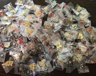 May include: A large collection of colorful enamel pins in individual plastic bags. The pins feature various animal and cartoon characters, including bears, butterflies, and birds.