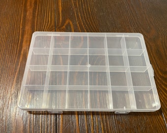 May include: A clear plastic storage box with 16 compartments. The box is empty and has a rectangular shape.