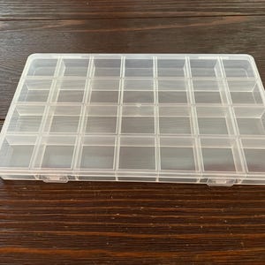May include: A clear plastic organizer with a rectangular shape and multiple small, square compartments. The organizer has a hinged lid and is designed for storing small items like beads, jewelry, or craft supplies. The container is sitting on a dark wooden surface.