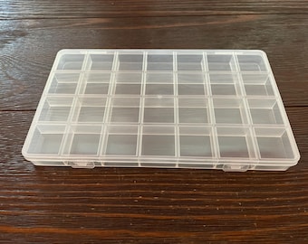 May include: A clear plastic organizer with a rectangular shape and multiple small, square compartments. The organizer has a hinged lid and is designed for storing small items like beads, jewelry, or craft supplies. The container is sitting on a dark wooden surface.
