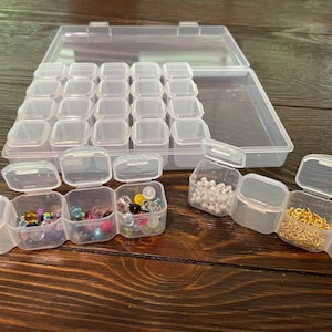 May include: A clear plastic organizer with multiple small compartments, some filled with colorful beads and gold-colored chain links. The organizer is open, revealing rows of empty compartments, and sits on a wooden surface.