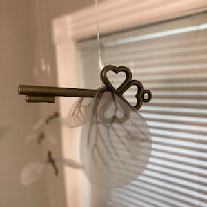 May include: A gold key with a heart-shaped design hangs from a white string. The key is attached to a pair of white, feathery wings.