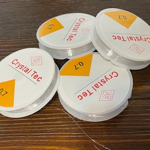 May include: Four spools of clear elastic string, each with a white label that says "Crystal Tec" and "0.7" in red and yellow lettering.