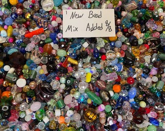May include: A large collection of assorted loose beads in various shapes, sizes, and colours including blue, red, green, yellow, and clear glass. A white rectangular card is placed on top with the handwritten text New Bead Mix Added 8/8.
