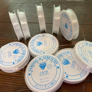 以下が含まれることがあります： 透明で伸縮性のあるビーズコードのスプールとストランド。スプールには、日本語のテキストとダイヤモンドのグラフィックが入った白と青のラベルが付いています。ラベルのテキストは「Strong & Stretchy」と「Crystalline」です。