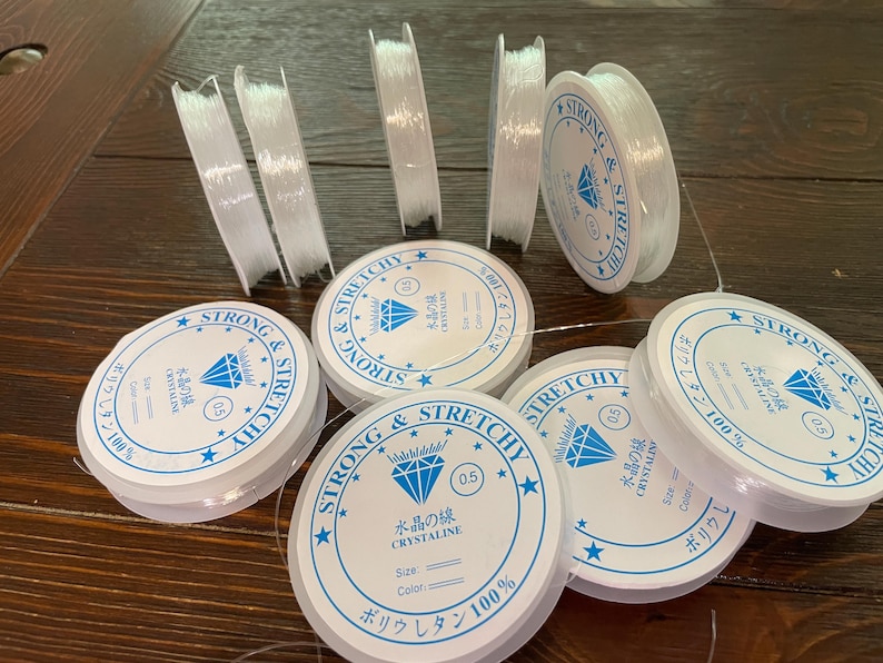 以下が含まれることがあります： 透明で伸縮性のあるビーズコードのスプールとストランド。スプールには、日本語のテキストとダイヤモンドのグラフィックが入った白と青のラベルが付いています。ラベルのテキストは「Strong & Stretchy」と「Crystalline」です。