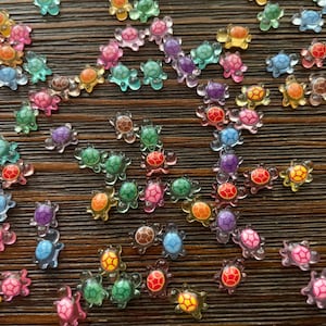 May include: A collection of small, colorful turtle-shaped embellishments. The turtles are translucent and come in various colors, including pink, blue, green, orange, and purple. They are scattered on a brown, textured surface.