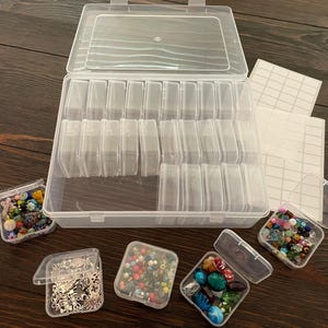 May include: A clear plastic storage organizer box featuring multiple small, removable individual containers inside. The set includes several small square cases filled with assorted colorful glass beads and silver metal charms, plus blank white labels.