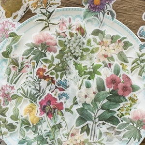 Puede incluir: Una colección de pegatinas florales con diversas flores y follaje en tonos rosa, morado, amarillo y verde. Las pegatinas están dispuestas sobre un plato blanco con un borde azul, creando una estética vintage.