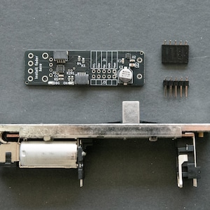 May include: A black printed circuit board with white text reading Install fader here, a silver motorized fader component, and two small black pin header connectors. The circuit board features various electronic components and solder points.