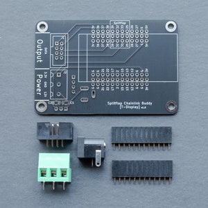 May include: A black circuit board labeled "Splitflap Chainlink Buddy [T-Display] v1.0" with various connectors and pin headers. The board has text indicating power and output connections, along with multiple pin designations. Several black and green connectors are arranged around the board.