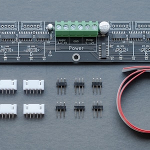 May include: A black circuit board with various electronic components, including chips, connectors, and a green terminal block. Additional components include white and black connectors, and a red and white wire. The board has text indicating "Input", "Output", and "Power".
