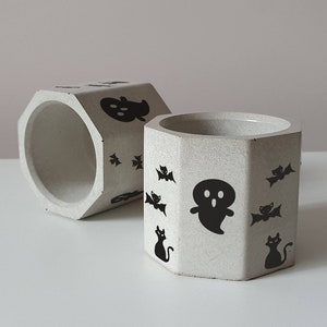 May include: Two white hexagonal concrete planters with black Halloween-themed designs. One planter features a ghost, bats, and black cats. The other planter features a ghost and bats.