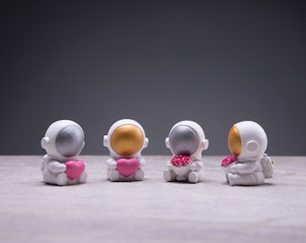 May include: Four white astronaut figurines with gold and silver helmets. Two are holding pink hearts and two are holding pink flowers.