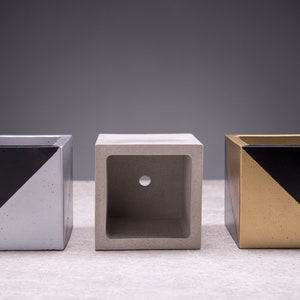 Può includere: Tre vasi in cemento con disegni geometrici in bianco e nero. Il vaso a sinistra è argentato, quello al centro è grigio e quello a destra è dorato.