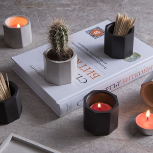 Puede incluir: Un conjunto de seis portavelas y portapalillos hexagonales de hormigón. Los soportes son de color negro y gris y presentan una variedad de artículos, como velas, palillos de dientes y una pequeña planta de cactus.