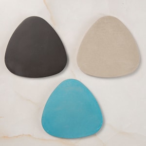 Puede incluir: Tres posavasos triangulares de cerámica en negro, beige y turquesa.