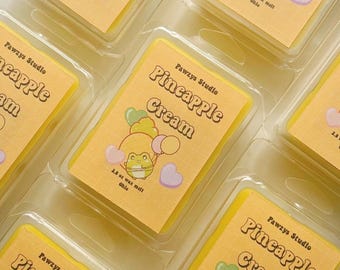 May include: Close-up of several yellow wax melts in clear plastic packaging. Each melt has a label with the text "Pawzys Studio Pineapple Cream" and a cartoon frog holding balloons. The melts are 2.6 oz each.