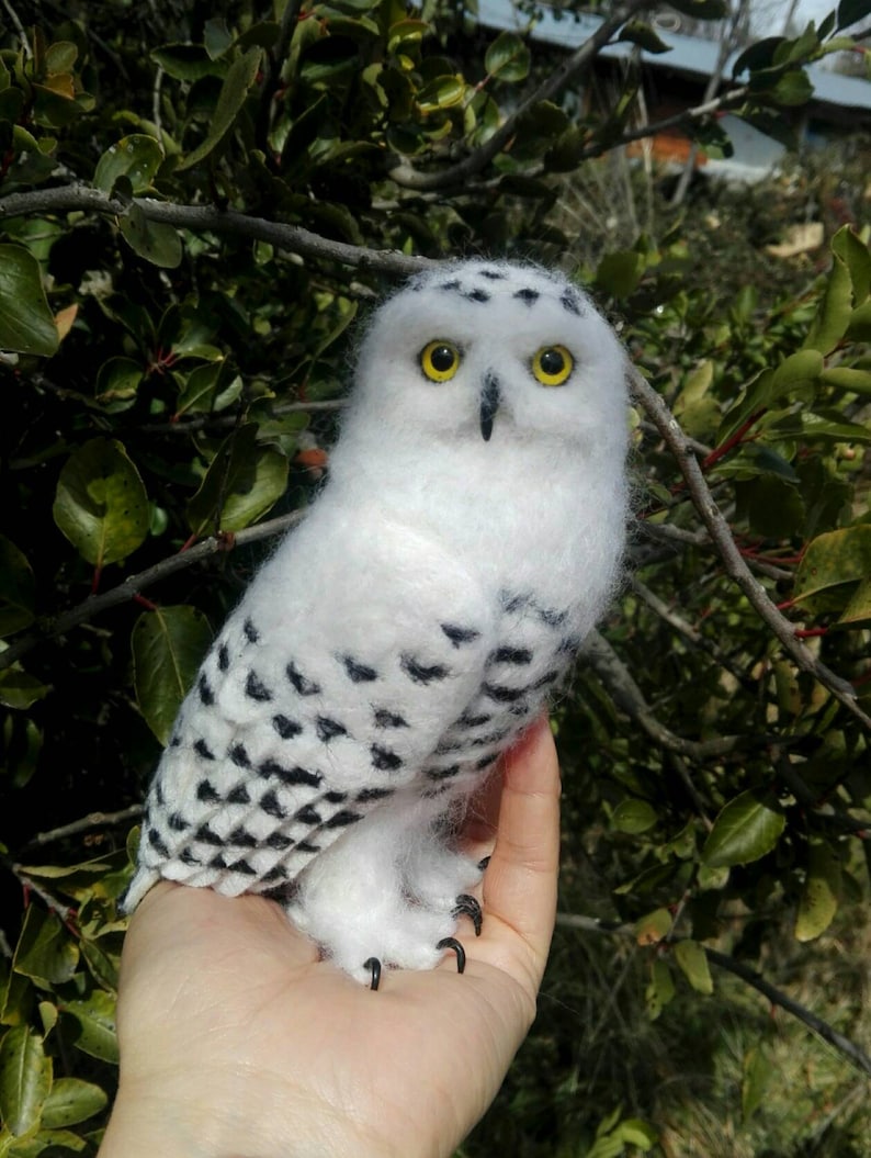 May include: A white and black snowy owl with yellow eyes is perched on a hand. The owl is made of felt and has a realistic appearance.