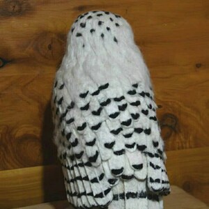 May include: A white and black needle felted snowy owl with black eyes. The owl is standing on a wooden surface.