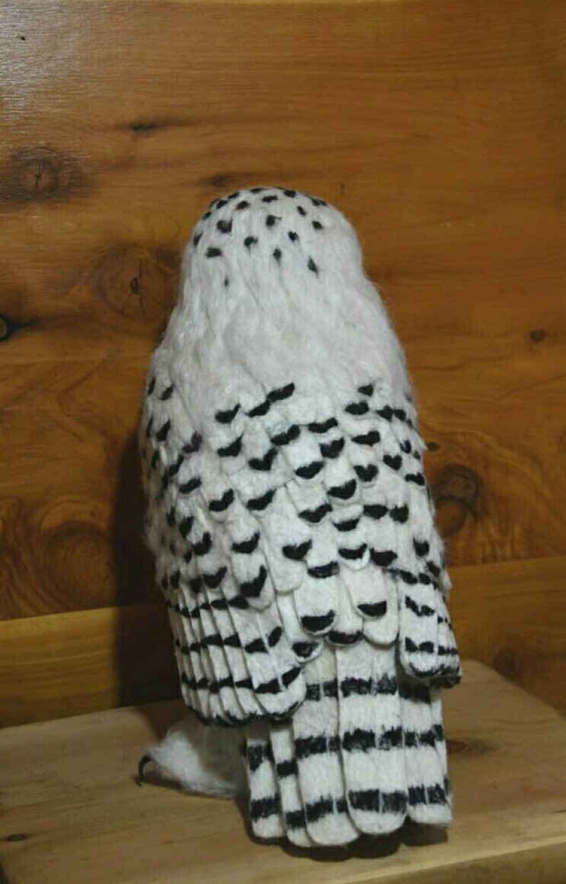 May include: A white and black needle felted snowy owl with black eyes. The owl is standing on a wooden surface.