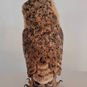 May include: A brown and white needle felted owl with black eyes and talons. The owl is standing on a white surface.