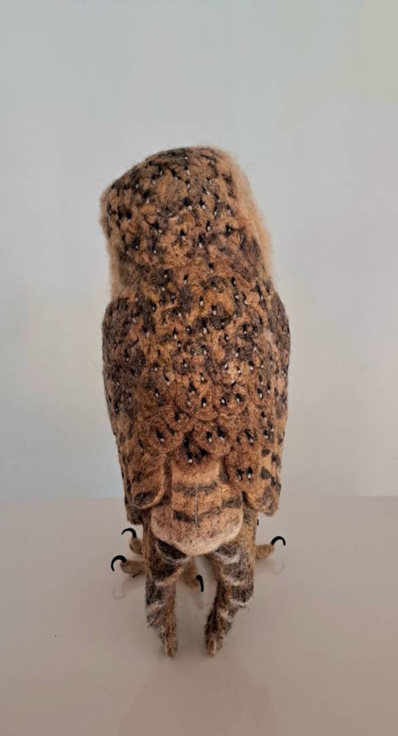 May include: A brown and white needle felted owl with black eyes and talons. The owl is standing on a white surface.