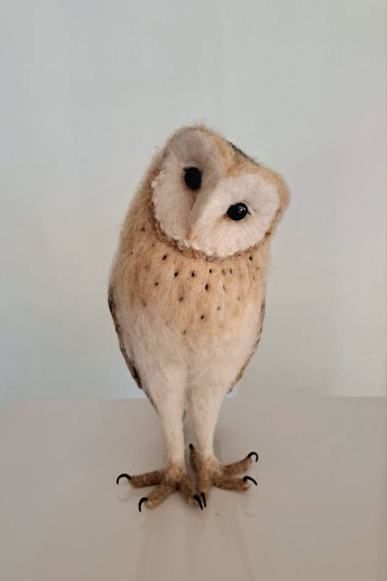 May include: A needle felted barn owl figurine with white and brown feathers. The owl is standing on a white surface and has its head tilted to the side.