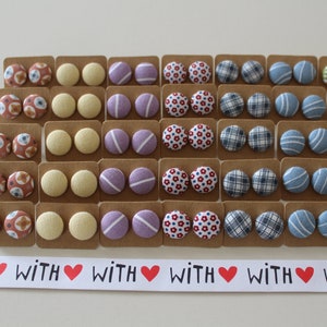May include: A set of 24 pairs of fabric button earrings. The earrings are displayed on small cards with the word "WITH" repeated on a white banner below. The earrings feature a variety of patterns and colors, including red, blue, yellow, green, and purple.