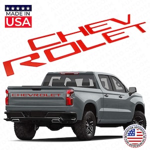 May include: Red Chevrolet truck decals spelling out CHEVROLET. The decals are shown above a gray pickup truck. The words "MADE IN USA" are in a red box in the upper left corner. The truck has black tires and a black bumper.
