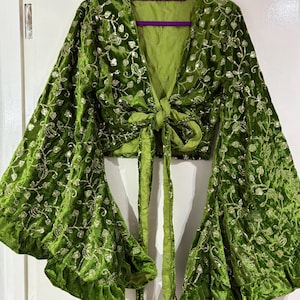 May include: A vibrant green velvet wrap top featuring long, wide bell sleeves and a deep V-neckline. The fabric is adorned with an intricate, shimmering gold floral embroidery pattern throughout, and the garment ties at the front waist.