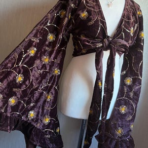 May include: A dark brown velvet wrap top featuring long, wide bell sleeves and a tie-front closure. The fabric is decorated with an embroidered pattern of gold and yellow floral motifs, accented with shimmering sequins throughout the design.