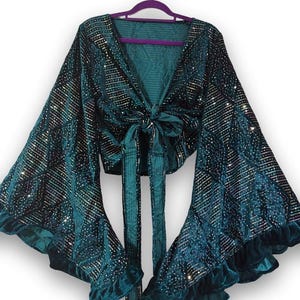 May include: A deep teal velvet wrap top featuring long, wide bell sleeves with ruffled cuffs. The fabric is adorned with a repeating diamond pattern composed of small, reflective silver sequins. The garment includes a front tie closure.