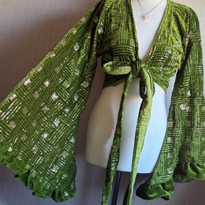 May include: A long-sleeved olive green velvet wrap top featuring a deep V-neckline and a front tie closure. The fabric is adorned with a geometric pattern of silver sequins and metallic thread, complemented by wide bell sleeves with ruffled cuffs.