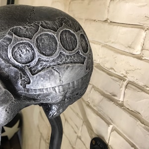May include: A silver skull sculpture with a brass knuckle design on the top of the head. The skull is mounted on a black metal stand.