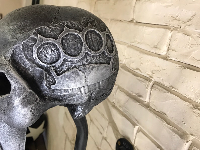 May include: A silver skull sculpture with a brass knuckle design on the top of the head. The skull is mounted on a black metal stand.