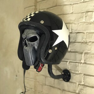 May include: A black motorcycle helmet with a white star and chequered pattern on the side. The helmet has a skull-shaped face mask and is mounted on a black metal wall hook.