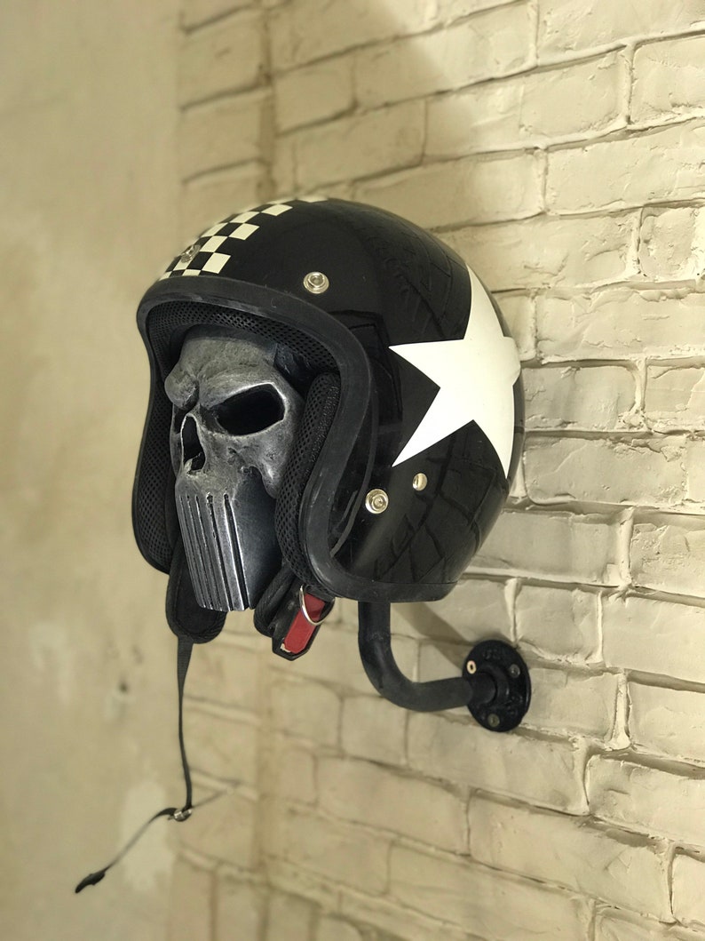 May include: A black motorcycle helmet with a white star and chequered pattern on the side. The helmet has a skull-shaped face mask and is mounted on a black metal wall hook.