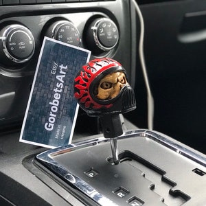 Peut inclure: Un pommeau de levier de vitesse en forme de crâne rouge et noir avec des flammes peintes sur le côté. Le pommeau est fixé au levier de vitesse d'une voiture. Une carte de visite pour "GorobetsArt" est visible en arrière-plan.