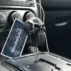 May include: A black and grey skull-shaped gear shift knob with a detailed design. The knob is attached to a silver gear shift lever in a car interior. A business card with the text "GorobetsArt Etsy Made in Ukraine" is visible in the image.
