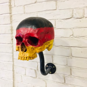Puede incluir: Un cráneo pintado de negro, rojo y amarillo con un tubo de metal negro unido a la parte posterior. El cráneo está montado en una pared de ladrillo blanco.