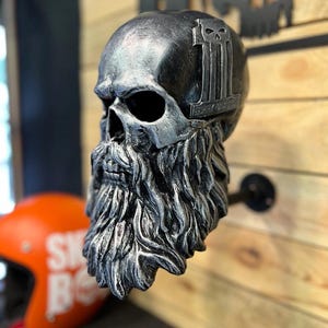 May include: A silver skull sculpture with a detailed, textured beard. The skull features a logo on top and is mounted on a wooden wall. An orange helmet is visible in the background. The sculpture is a decorative item.