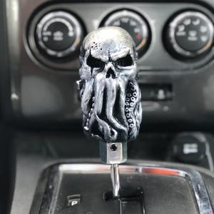 以下が含まれることがあります： タコの手のひらのデザインが施された銀色の頭蓋骨のシフトノブ。ノブは銀色のシフトレバーに取り付けられています。