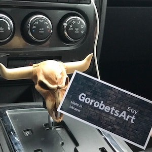 Op de afbeelding: Een witte stieren schedel versnellingspookknop met hoorns. De knop is bevestigd aan de versnellingspook van een auto. Een visitekaartje met de tekst "GorobetsArt Etsy Gemaakt in Oekraïne" is op de voorgrond.