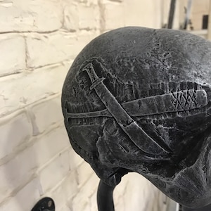 May include: A black skull wall hook with a textured surface and a design of crossed swords. The hook is attached to a black metal pipe.