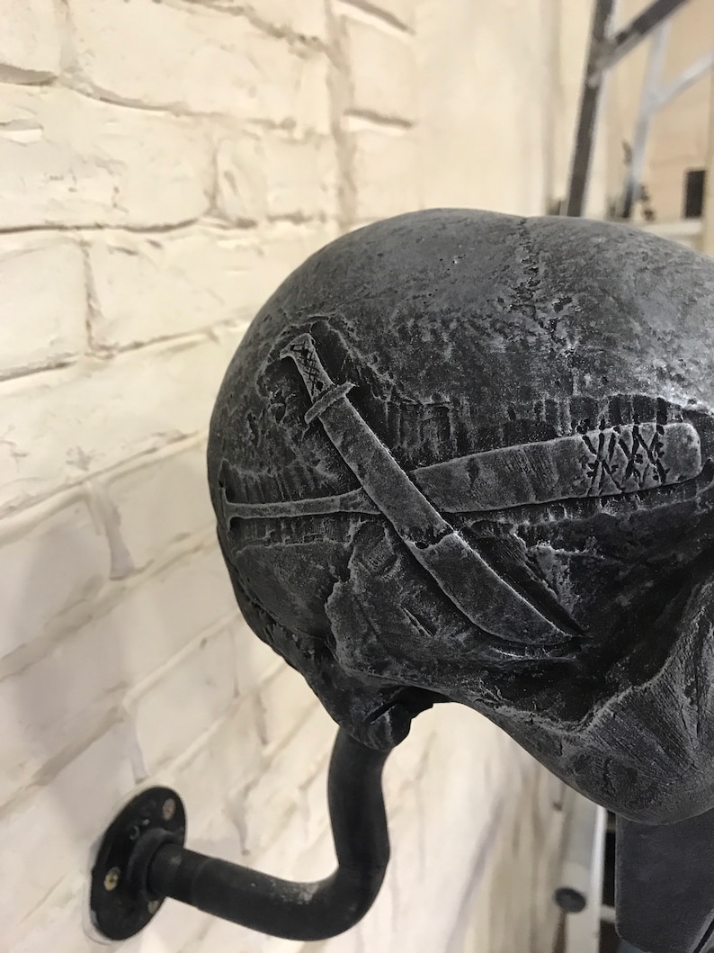 May include: A black skull wall hook with a textured surface and a design of crossed swords. The hook is attached to a black metal pipe.