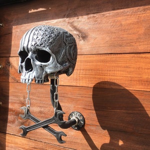 Op de afbeelding: Een zilveren schedel met een Route 66-ontwerp en een ketting die aan zijn kaak hangt. De schedel is gemonteerd op een metalen pijp met twee gekruiste moersleutels eronder. De achtergrond is een houten muur.