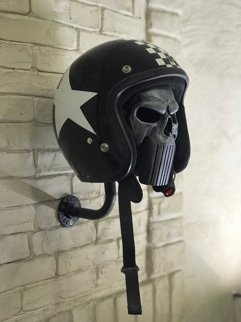 May include: A black motorcycle helmet with a white star and chequered pattern on the side. The helmet has a skull-shaped face mask with a black and white striped design. The helmet is mounted on a black metal wall mount.