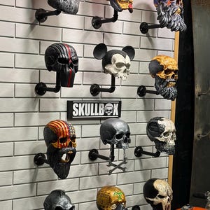 May include: A wall display featuring 12 skull-shaped figurines, each with a unique design and colour scheme. The skulls are mounted on black metal pipes and arranged in a grid pattern. The word "SKULLBOX" is displayed on a black sign in the centre of the wall.